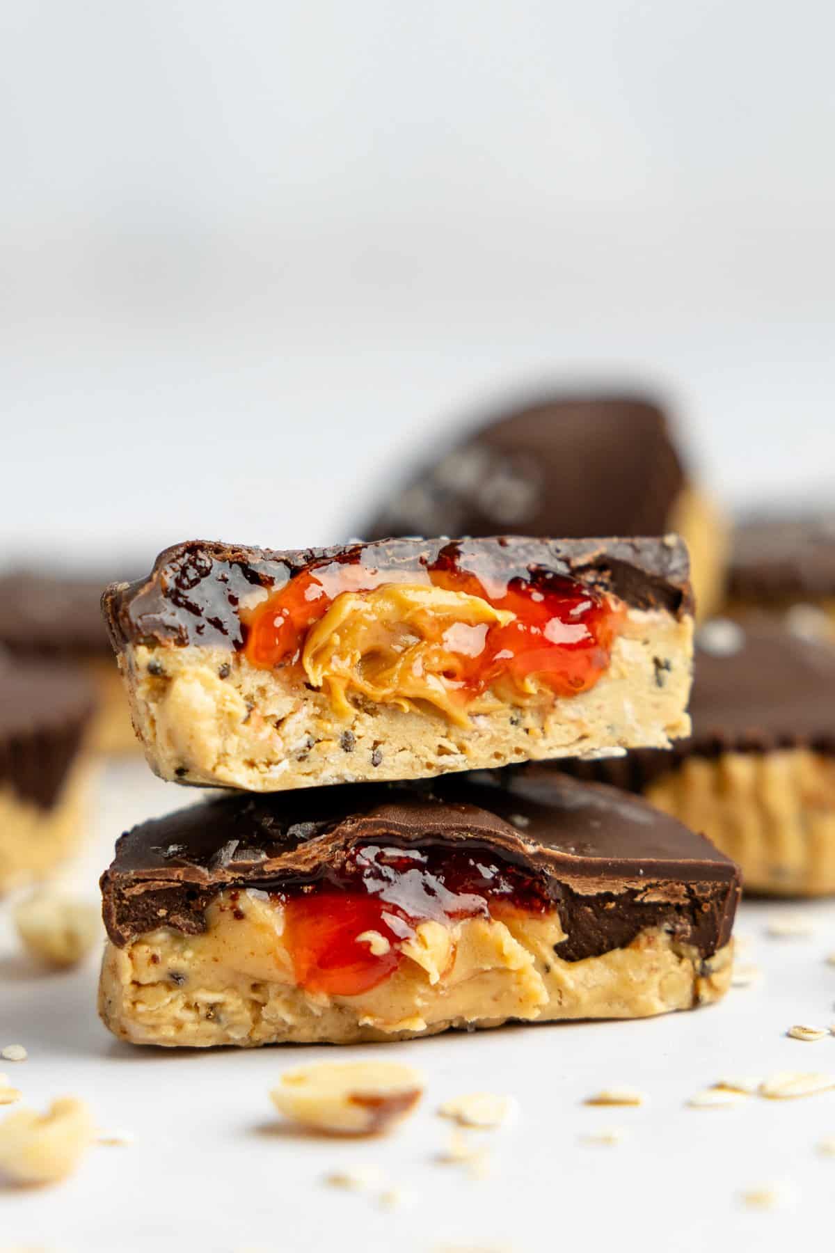 Two chocolate peanut butter cups, one cut in half, revealing peanut butter and jelly filling. Surrounded by more peanut butter cups and scattered oats on a white surface.