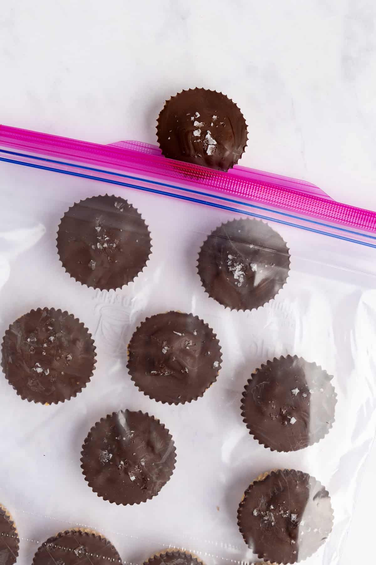 Chocolate cups with sea salt on top are inside a transparent resealable bag, with one cup placed above the bag.