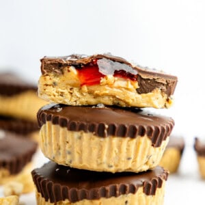 Three peanut butter and chocolate cups stacked, with a bite taken from the top one, revealing a red filling. Peanuts scattered around.