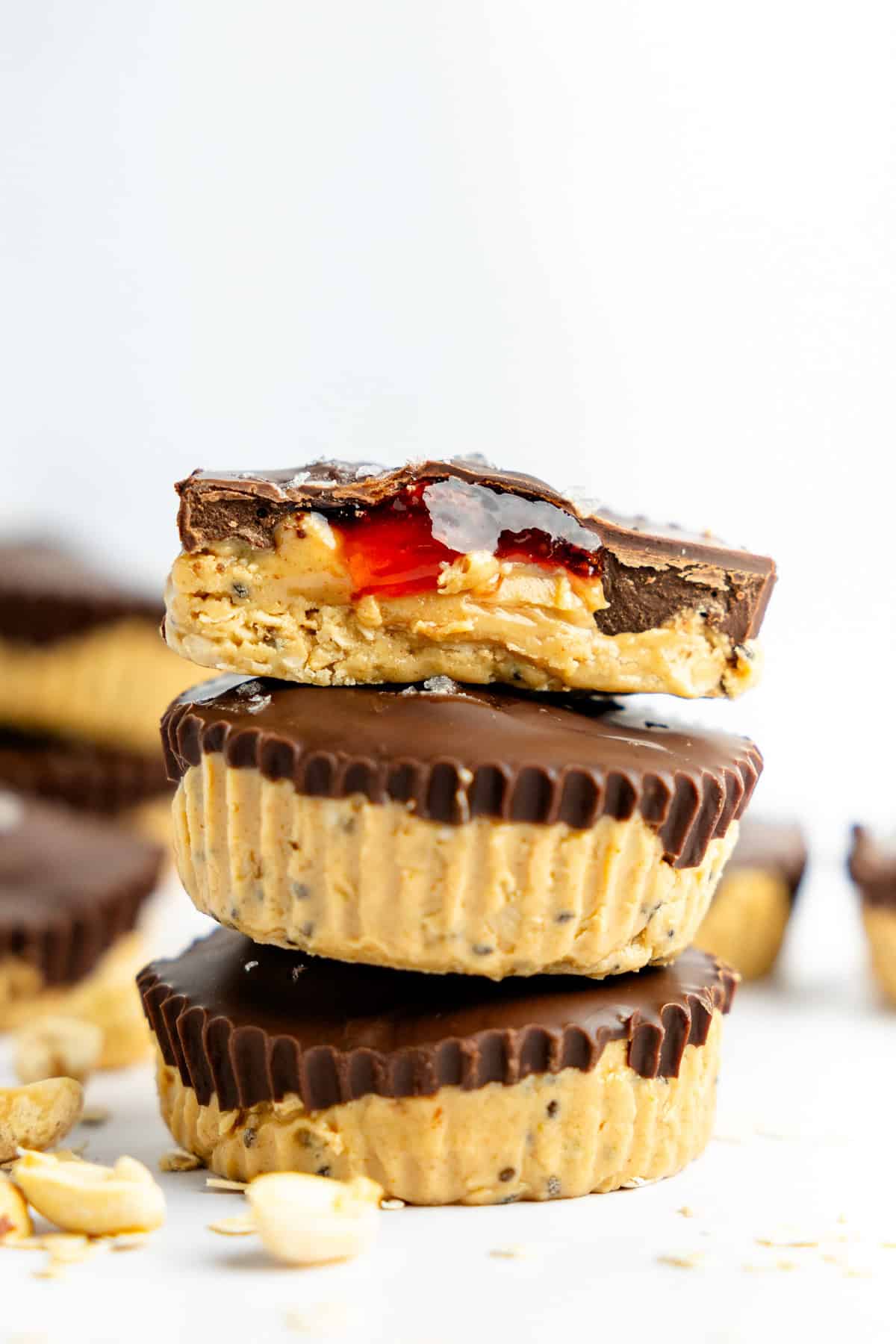 Three peanut butter and chocolate cups stacked, with a bite taken from the top one, revealing a red filling. Peanuts scattered around.