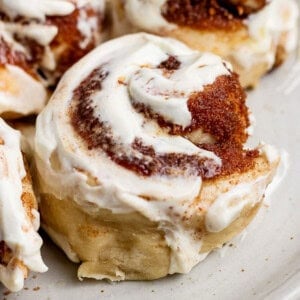 Close-up of a cinnamon roll with creamy white icing and swirls of cinnamon sugar on a light-colored plate.