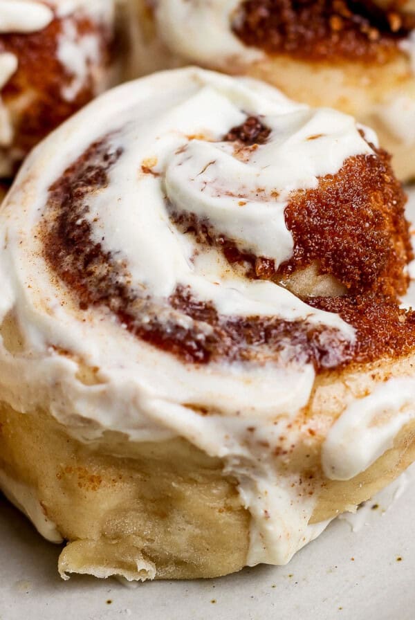 Close-up of a cinnamon roll with creamy white icing and swirls of cinnamon sugar on a light-colored plate.