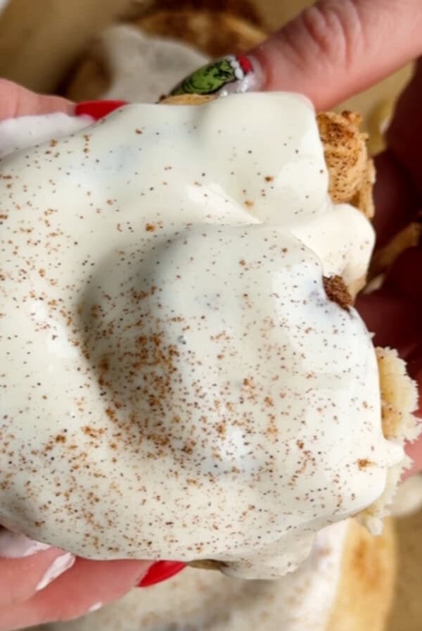 Hands holding a protein-infused cinnamon roll topped with cream cheese frosting and a sprinkle of cinnamon.