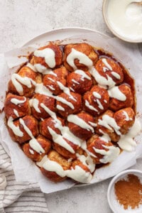 A round cinnamon pull-apart bread topped with cream cheese icing on parchment paper. Nearby are a bowl of icing and a dish of cinnamon.