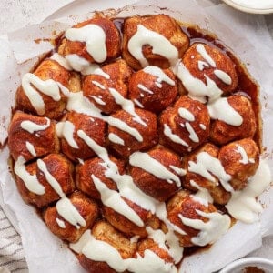 A round cinnamon pull-apart bread topped with cream cheese icing on parchment paper. Nearby are a bowl of icing and a dish of cinnamon.