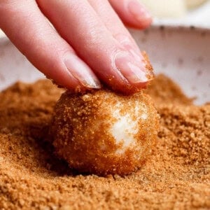 A hand rolls a dough ball in a bowl of brown sugar topping.