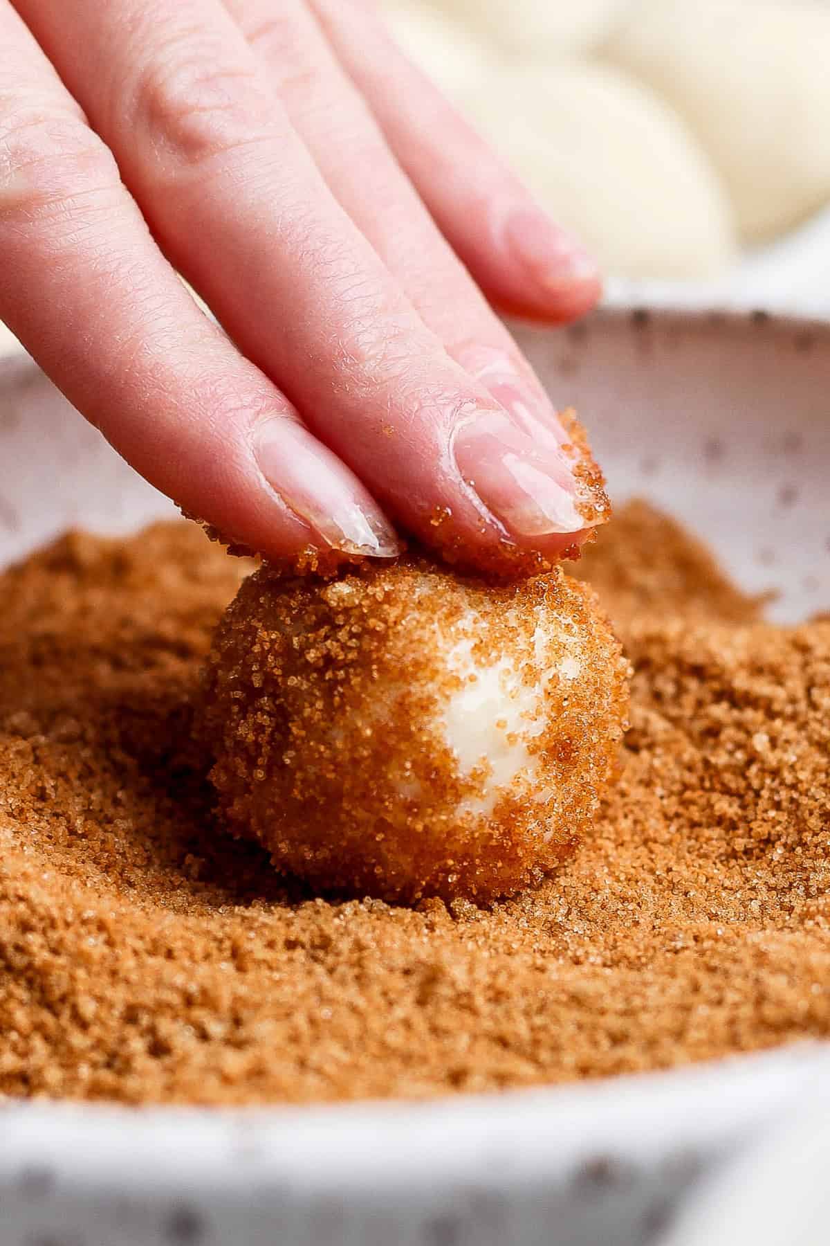 A hand rolls a dough ball in a bowl of brown sugar topping.