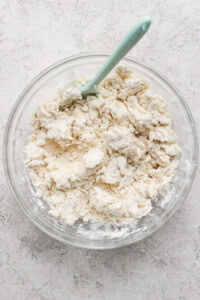 A clear bowl with a light blue spatula contains clumps of mixed flour and butter, forming a crumbly dough, on a textured gray surface.