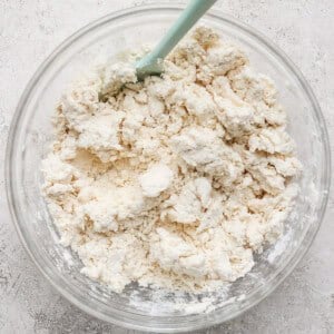 A clear bowl with a light blue spatula contains clumps of mixed flour and butter, forming a crumbly dough, on a textured gray surface.