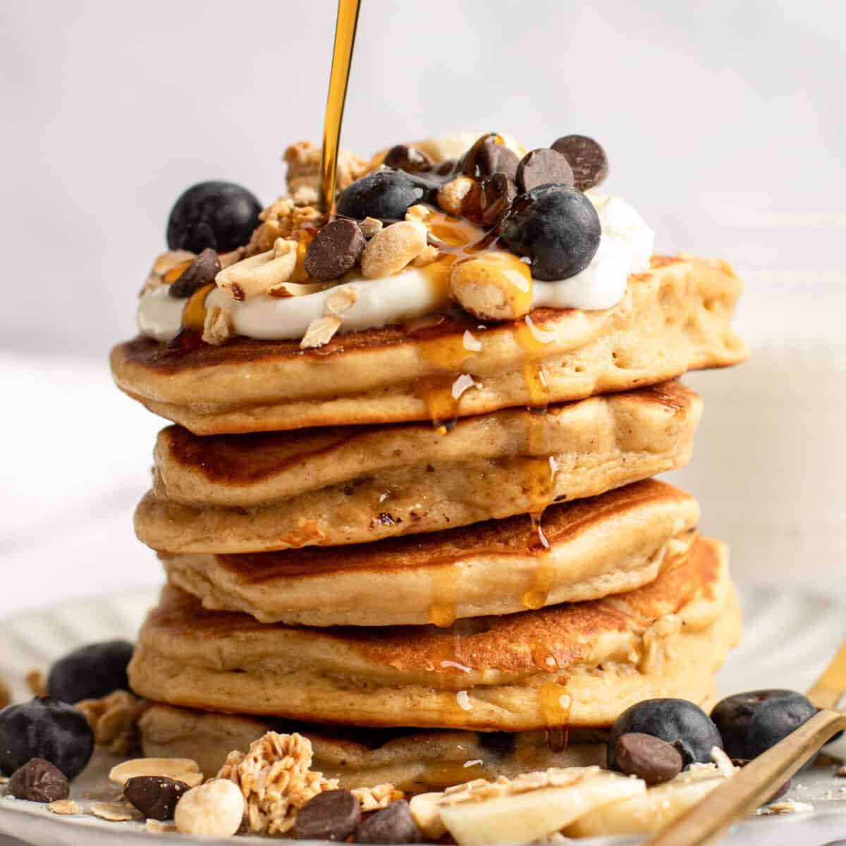 A stack of pancakes topped with blueberries, granola, yogurt, chocolate chips, and syrup being poured over them.