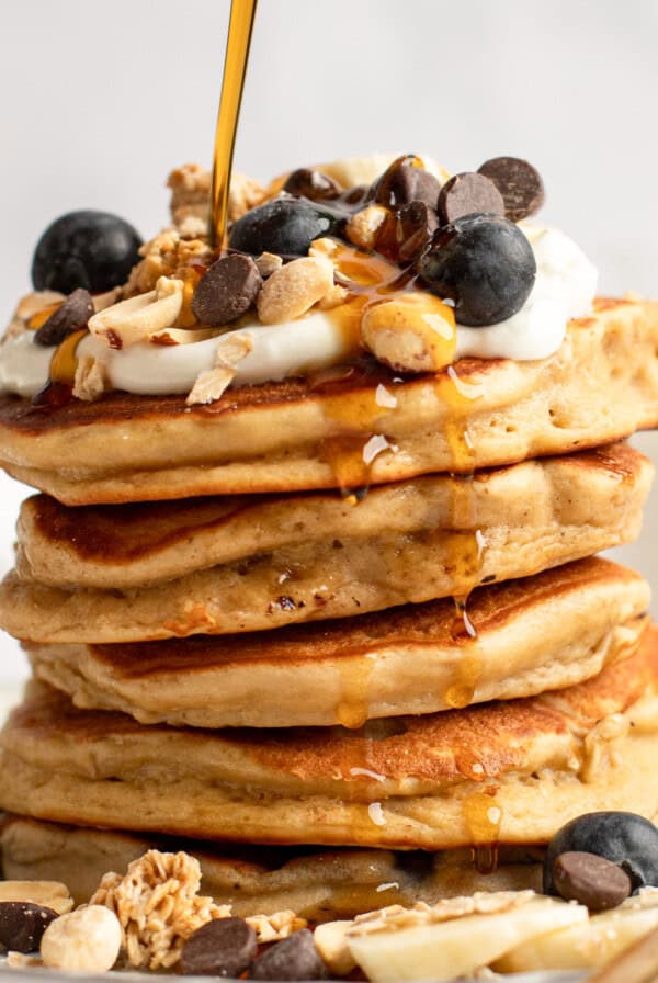 A stack of pancakes topped with blueberries, granola, yogurt, chocolate chips, and syrup being poured over them.