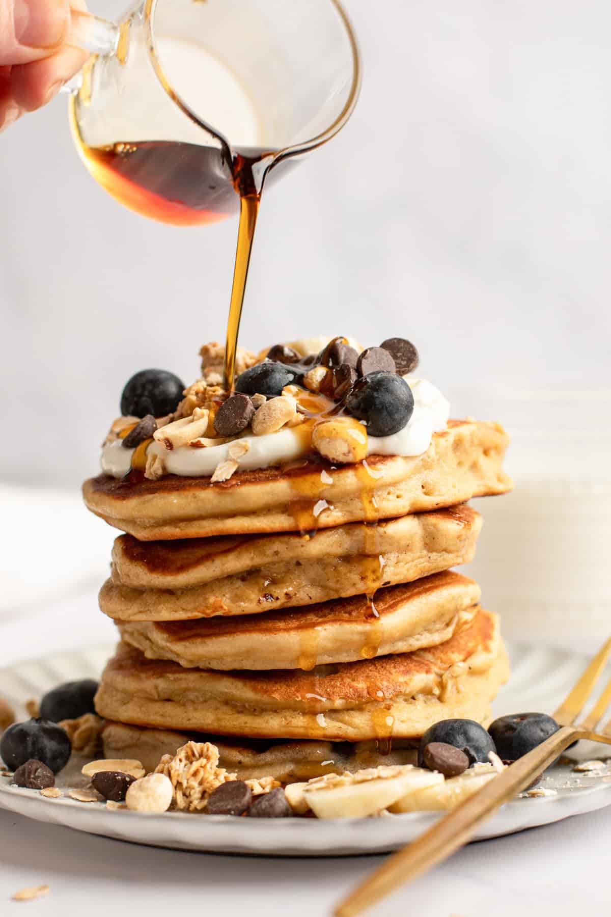 A stack of pancakes topped with blueberries, chocolate chips, granola, and nuts. Syrup is being poured over them.