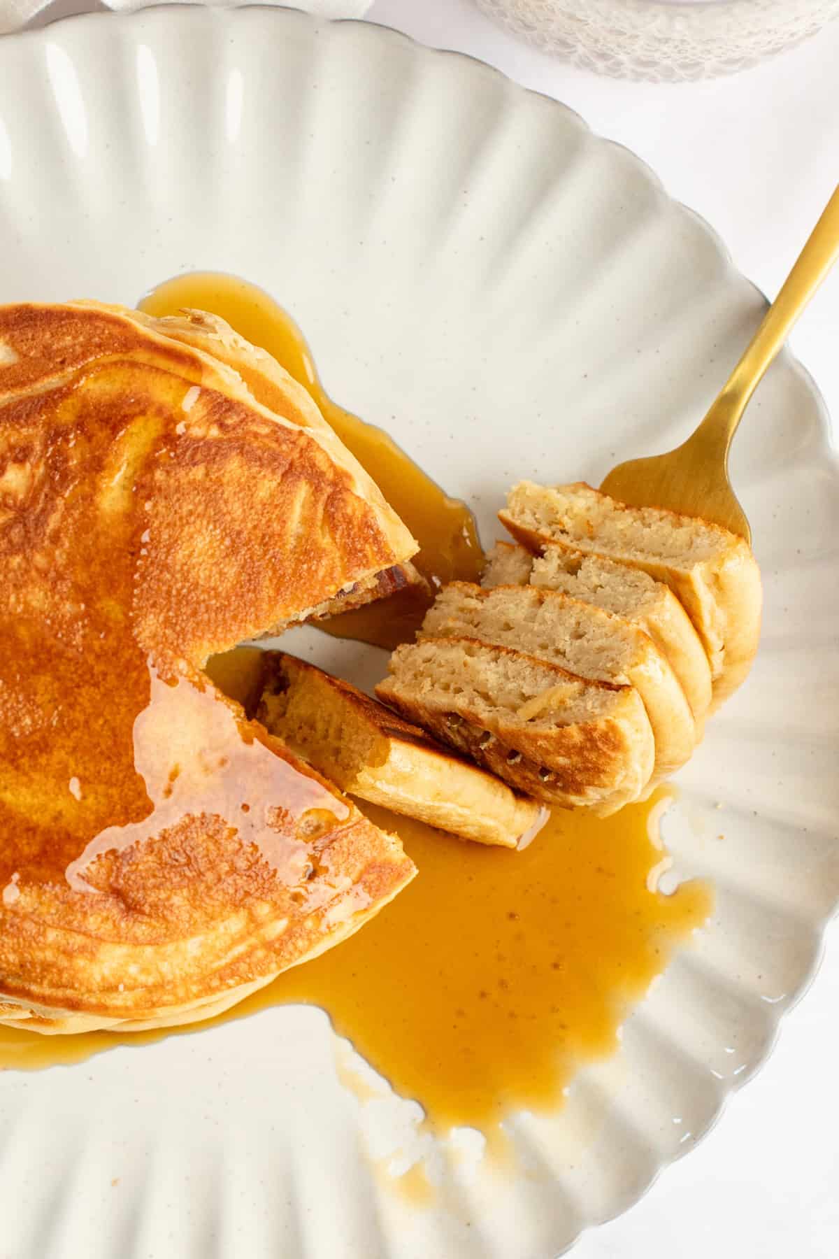 A plate of pancakes with syrup, partially eaten, sits on a white dish. A fork holds a cut portion of the pancakes.