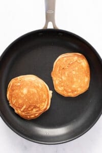 Two golden-brown pancakes cooking in a black non-stick frying pan on a white surface.