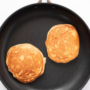 Two golden-brown pancakes cooking in a black non-stick frying pan on a white surface.