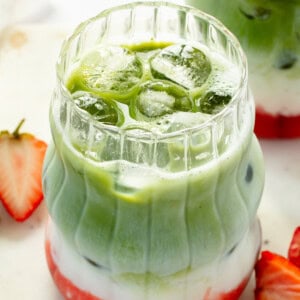 Layered matcha latte with ice, featuring green matcha, white milk, and red strawberry syrup. Served in a clear, textured glass with sliced strawberries on the side.