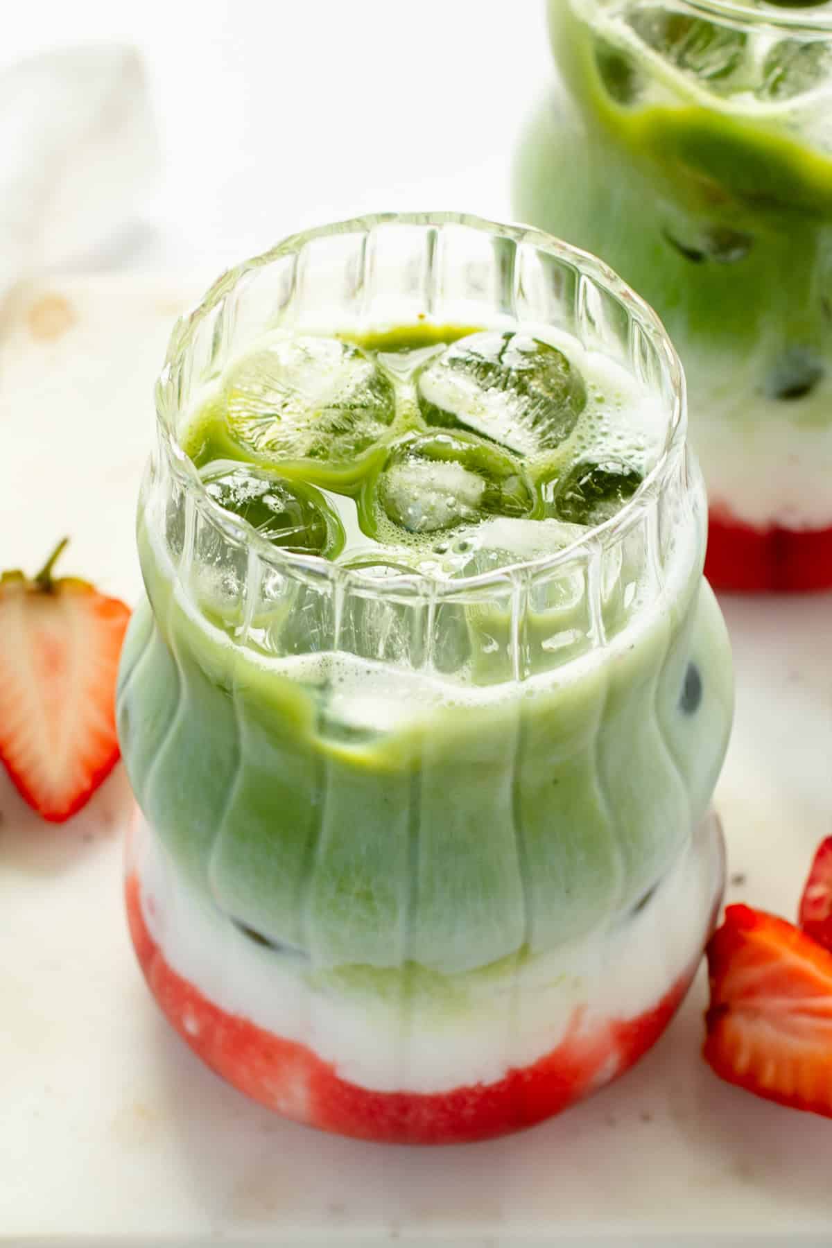 Layered matcha latte with ice, featuring green matcha, white milk, and red strawberry syrup. Served in a clear, textured glass with sliced strawberries on the side.