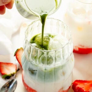 Milk being poured into a glass with strawberries and green liquid.