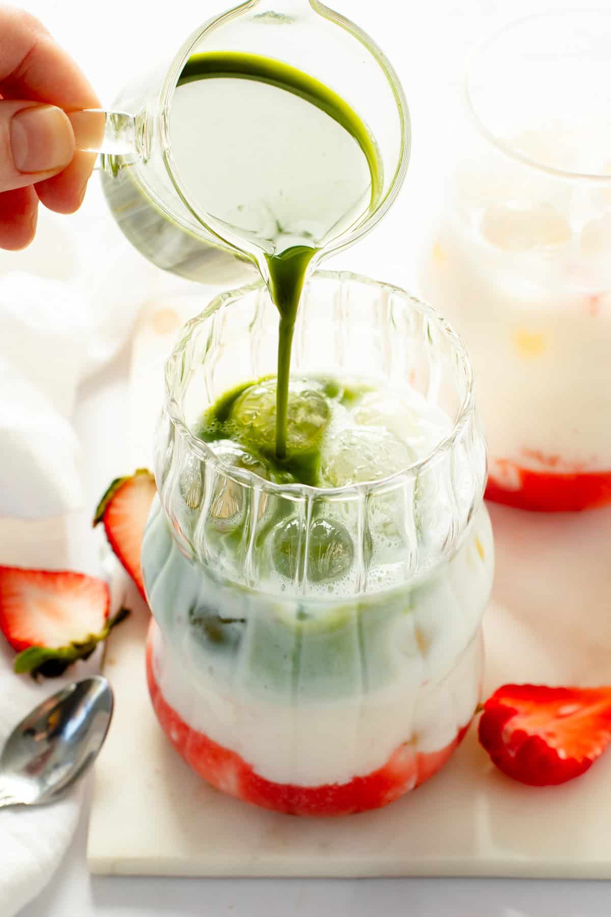 Milk being poured into a glass with strawberries and green liquid.