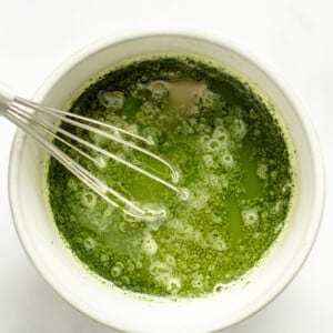 A whisk in a white bowl containing frothy green matcha tea against a white background.