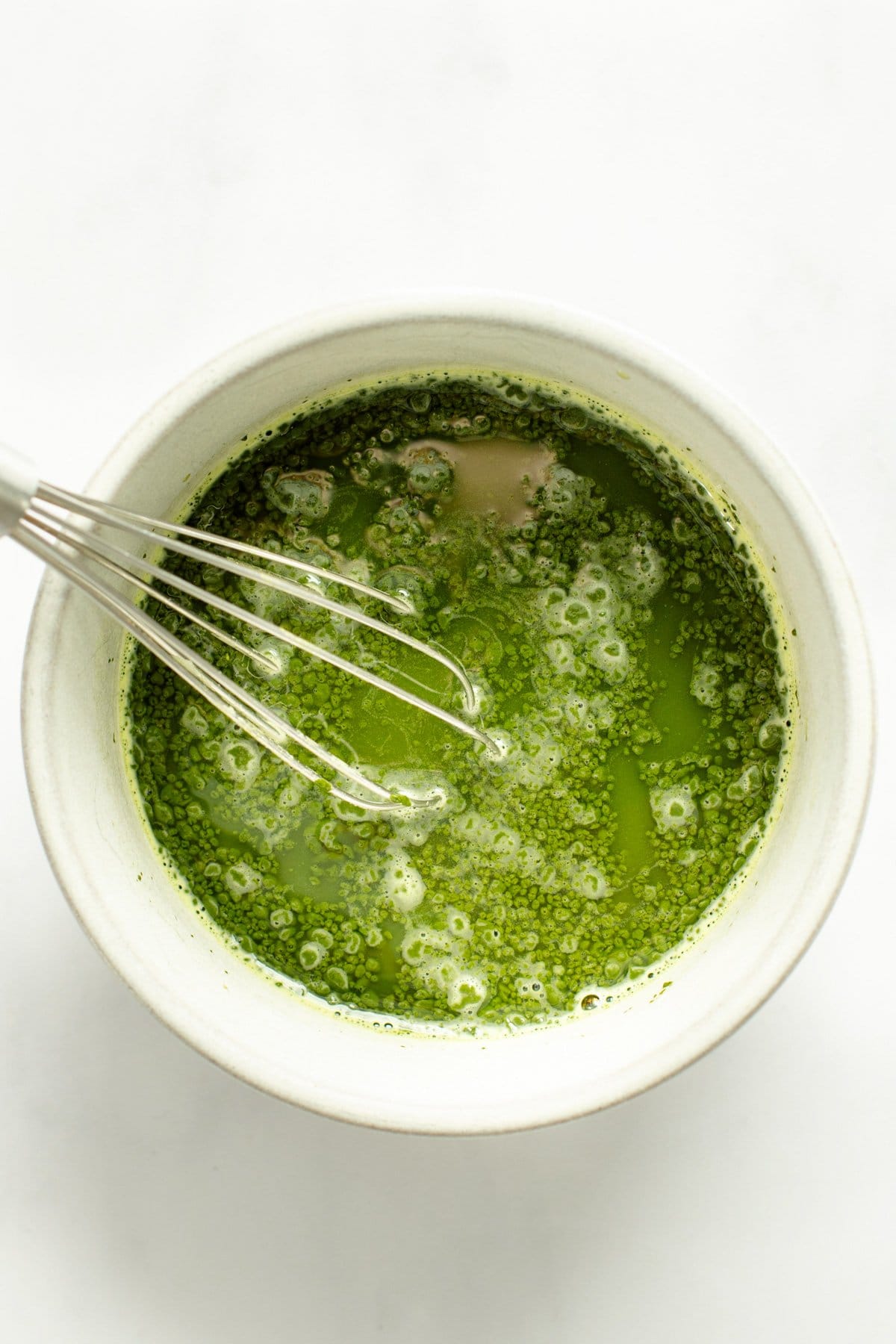 A whisk in a white bowl containing frothy green matcha tea against a white background.