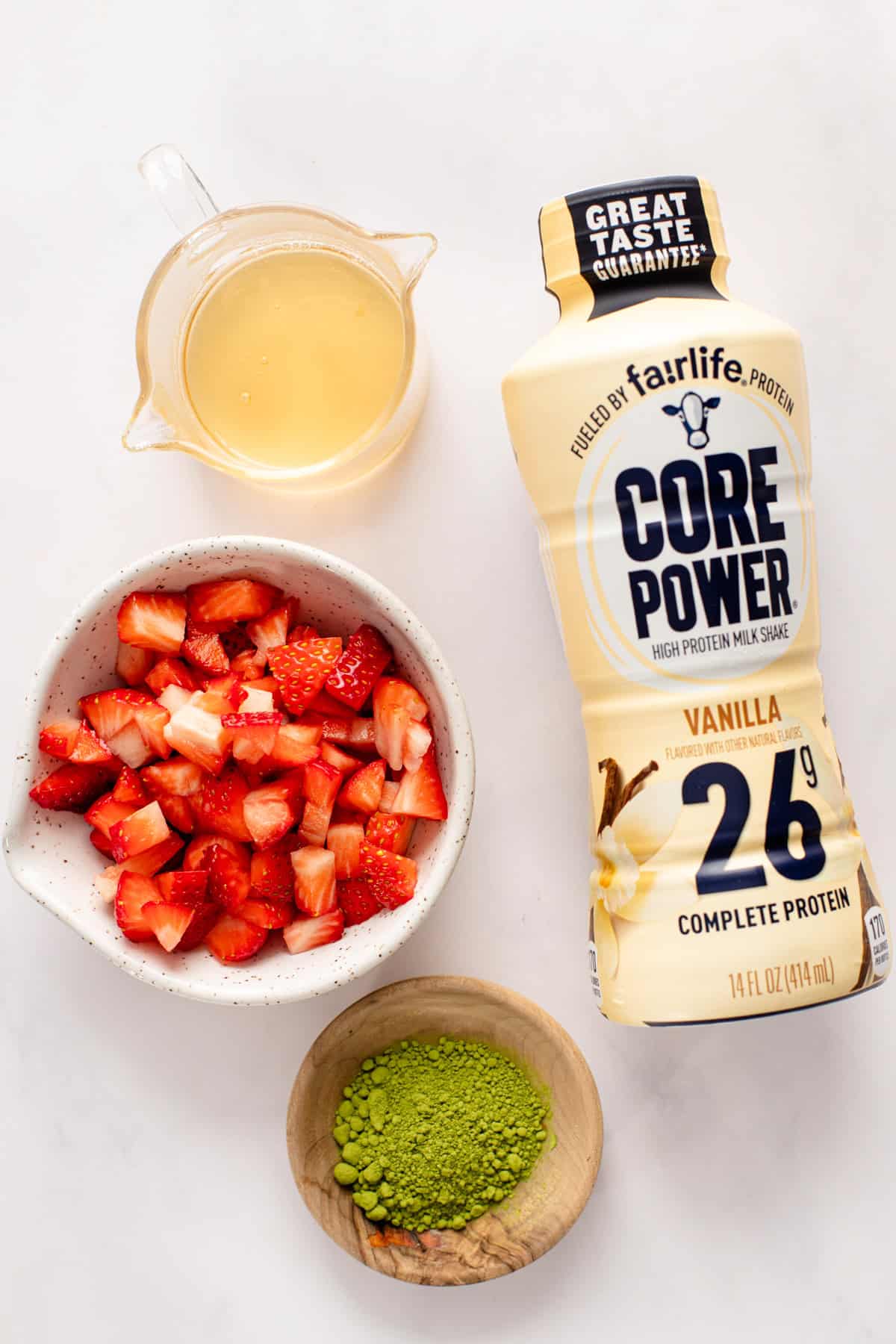 Vanilla protein milkshake bottle next to a small jug of liquid, a bowl of chopped strawberries, and a bowl of matcha powder on a white surface.