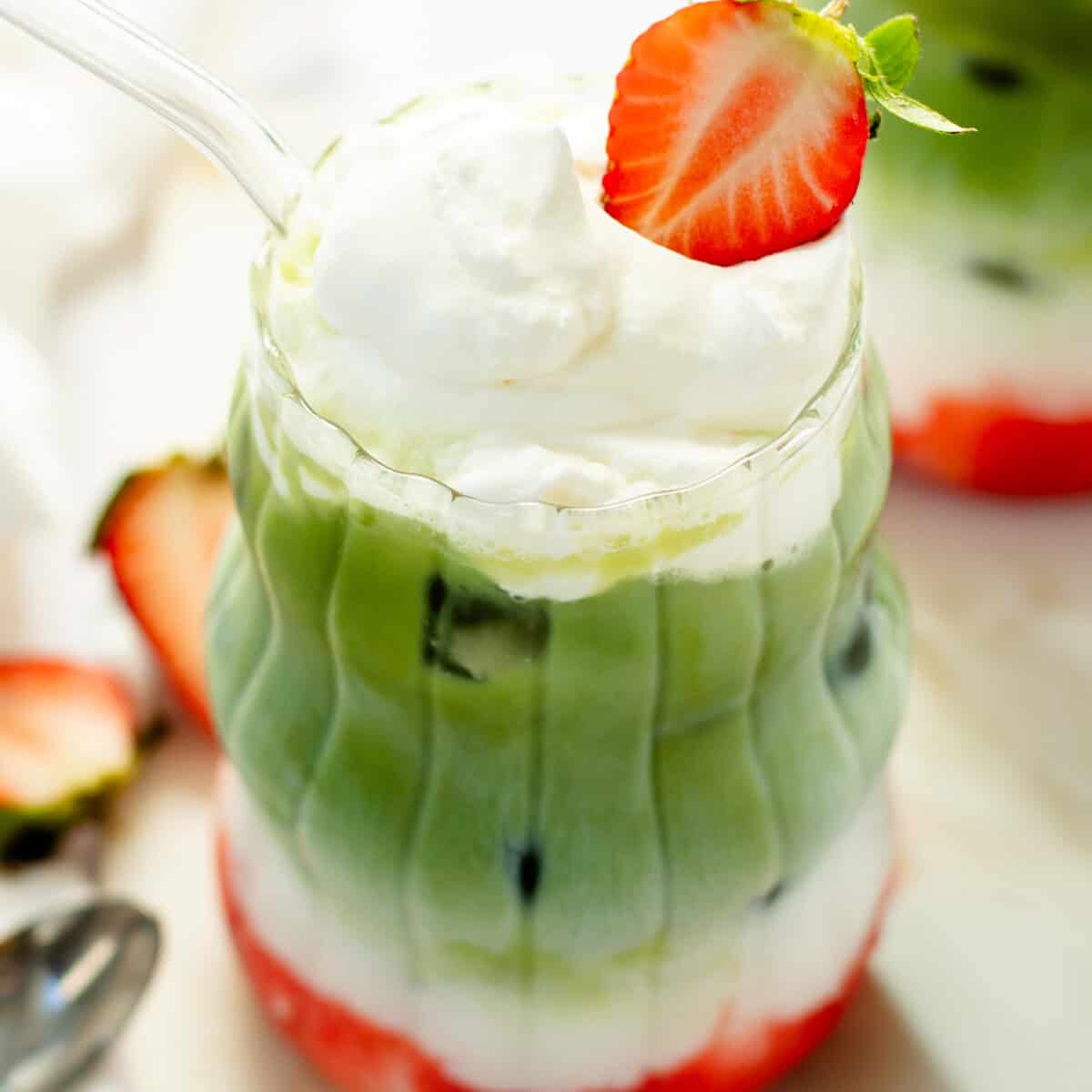 Layered drink with matcha green tea, milk, and strawberry puree, topped with whipped cream and half a strawberry in a clear glass with a straw.