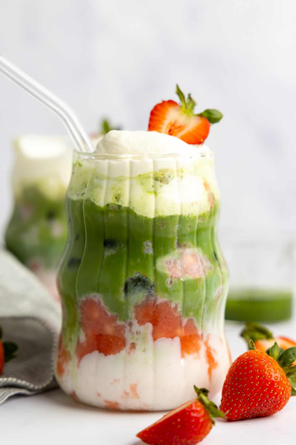 A layered drink featuring protein milk, green matcha, vibrant fruit pieces, and whipped cream in a clear glass. It's elegantly topped with a strawberry slice and accompanied by a transparent straw.