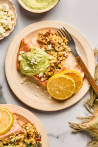 Plate with salmon topped with green sauce, alongside couscous, crumbled cheese, and lemon slices. Fork on the plate, napkin beside it.