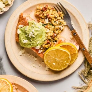 Plate with salmon topped with green sauce, alongside couscous, crumbled cheese, and lemon slices. Fork on the plate, napkin beside it.