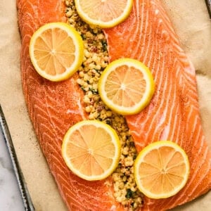 Raw salmon fillet stuffed with seasoned breadcrumbs and topped with lemon slices on parchment paper.