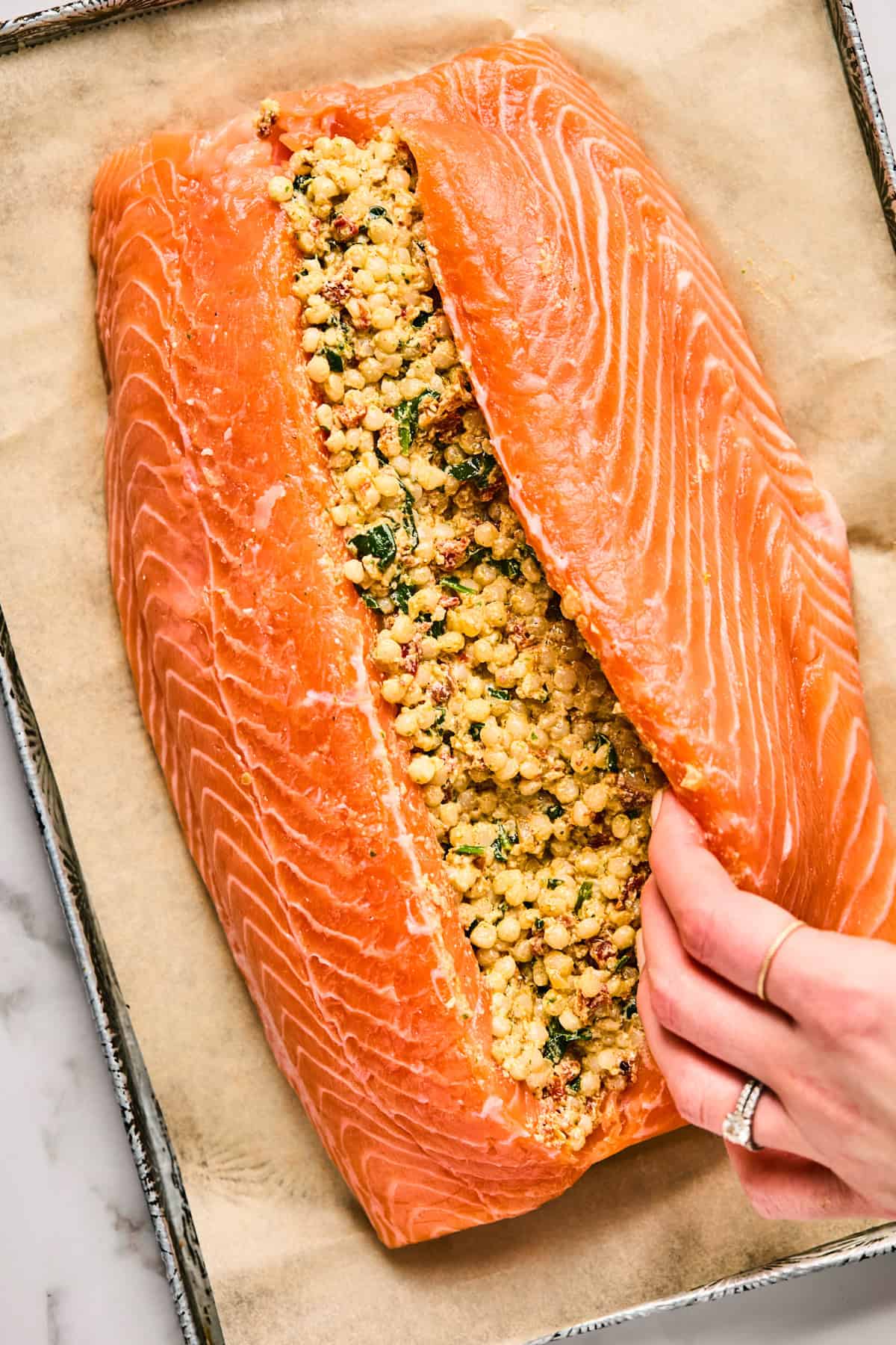 Raw salmon fillet is slit open and filled with a mixture of herbs and grains on a parchment-lined baking tray. A hand adjusts the fillet.