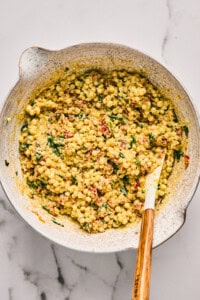 A bowl filled with a couscous salad mixed with herbs, spices, and sun-dried tomatoes on a marble surface. A wooden spoon rests in the bowl.