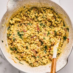 A bowl filled with a couscous salad mixed with herbs, spices, and sun-dried tomatoes on a marble surface. A wooden spoon rests in the bowl.