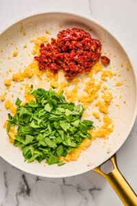 Chopped spinach, diced onions, and sun-dried tomatoes cooking in a frying pan on a marble surface.