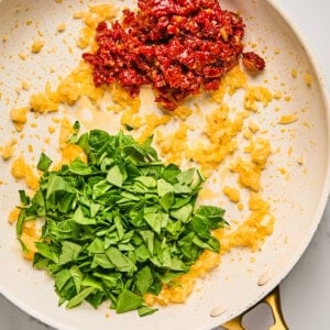 Chopped spinach, diced onions, and sun-dried tomatoes cooking in a frying pan on a marble surface.