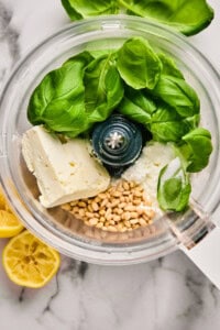 Food processor with basil leaves, cheese, pine nuts, and lemon halves on a marble surface.