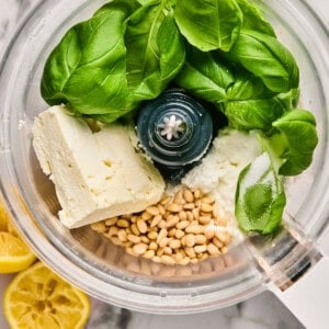 Food processor with basil leaves, cheese, pine nuts, and lemon halves on a marble surface.