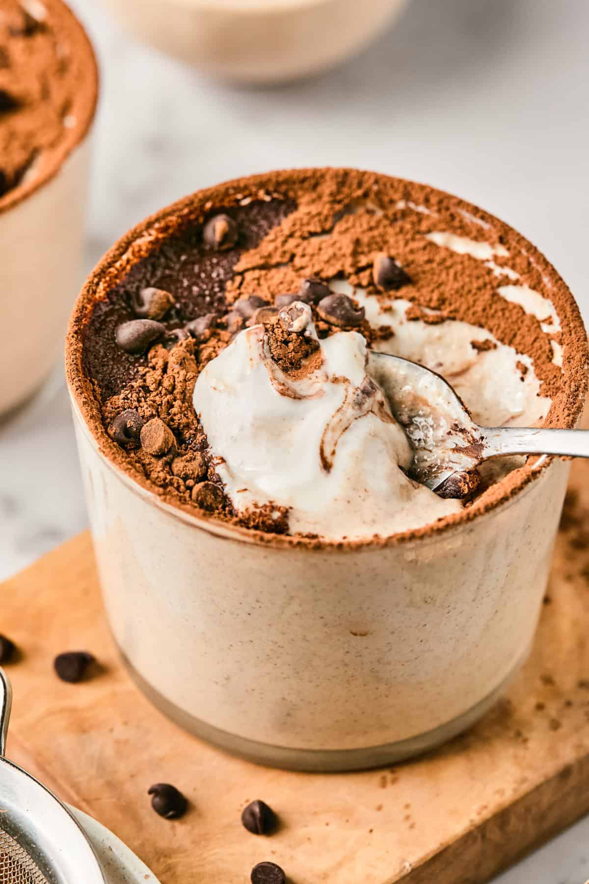 Tiramisu in a ceramic cup topped with cocoa powder and chocolate chips, with a spoon scooping a portion.