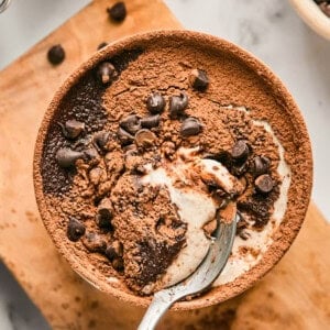 A bowl of chocolate dessert topped with cocoa powder and chocolate chips, partially eaten, with a spoon resting inside.