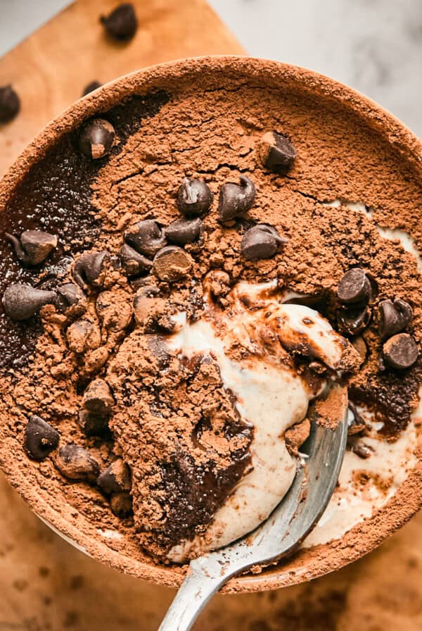 A bowl of chocolate dessert topped with cocoa powder and chocolate chips, partially eaten, with a spoon resting inside.