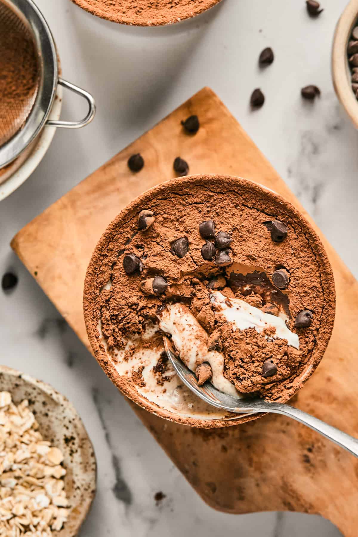 Bowl of tiramisu topped with cocoa powder and chocolate chips on a wooden board, next to a sieve, oats, and scattered chocolate chips. A spoon is scooping a portion from the bowl.