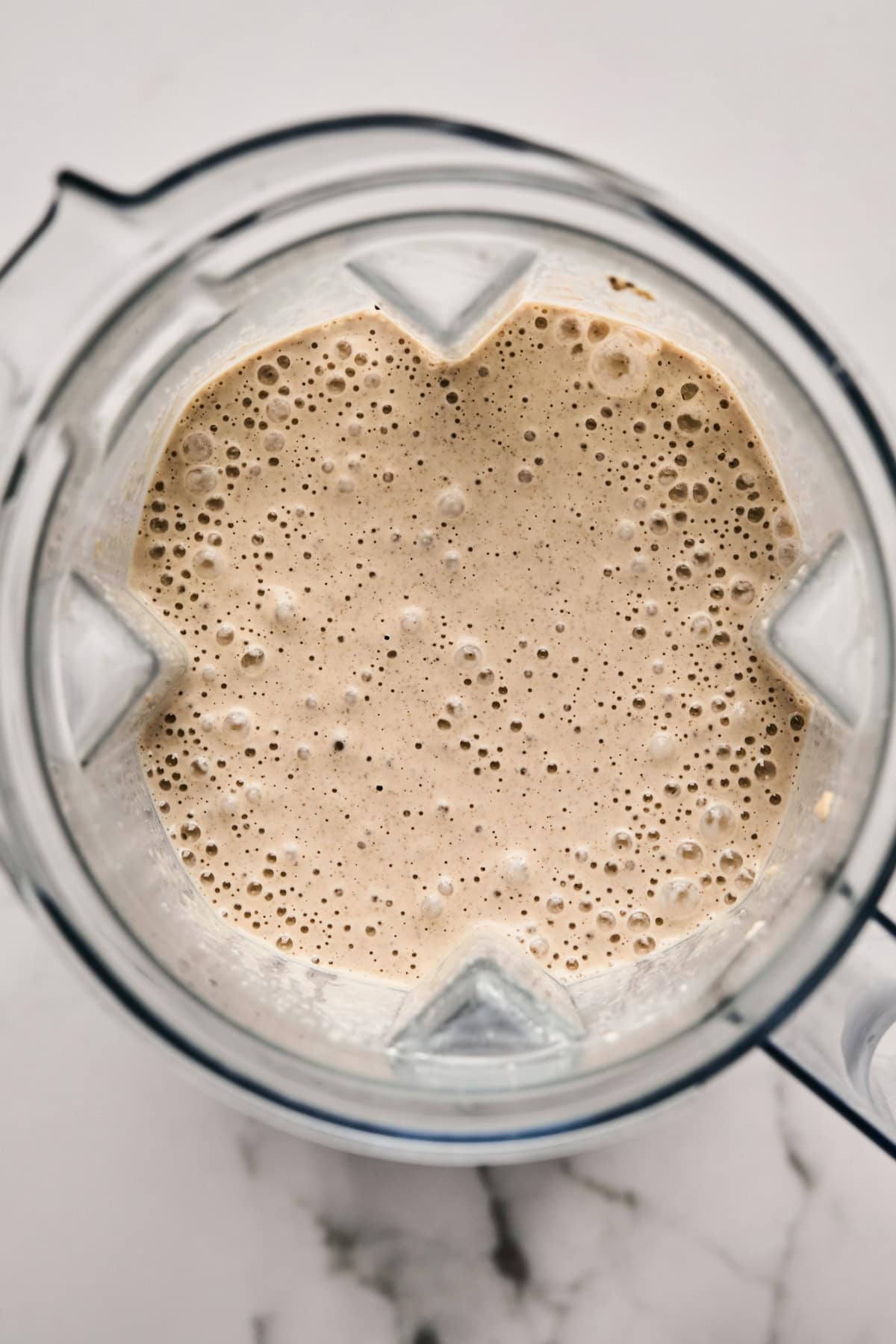 Top view of a blender filled with a frothy beige liquid, possibly a smoothie, on a white marble surface.