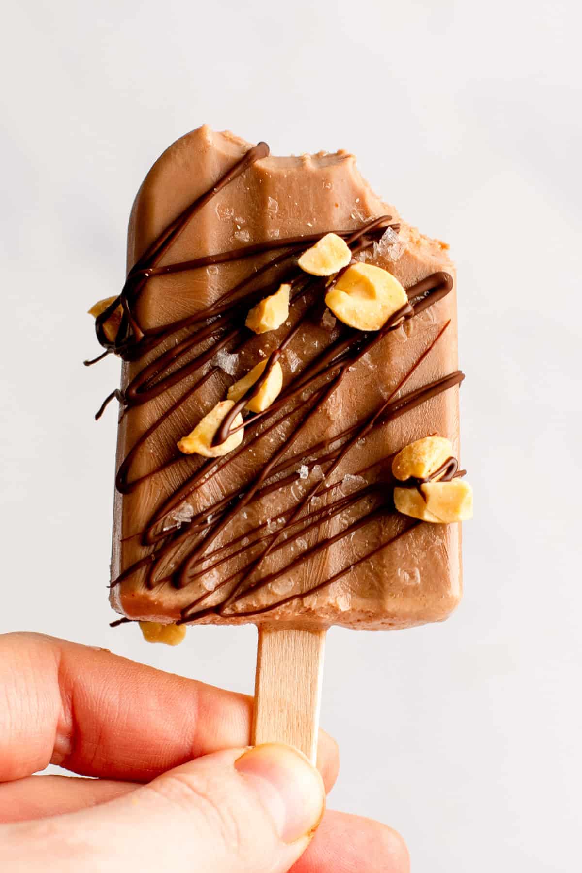 Person holding a chocolate popsicle with drizzled chocolate and peanut pieces, partially eaten.