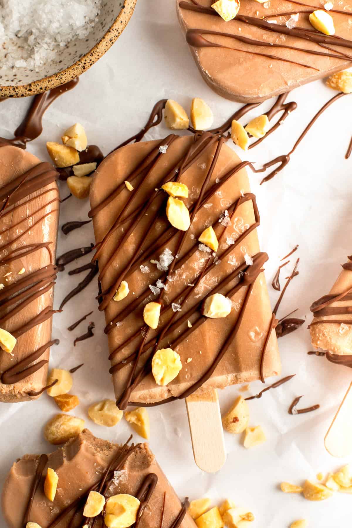 Chocolate popsicles drizzled with chocolate and sprinkled with chopped nuts and sea salt on a white surface.