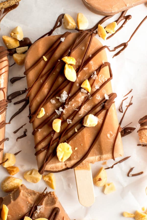 Close-up of a chocolate ice cream bar on a stick, drizzled with chocolate and sprinkled with chopped nuts and sea salt, on a white surface.