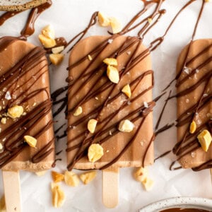 Three chocolate popsicles drizzled with dark chocolate and sprinkled with crushed nuts and sea salt are arranged on a parchment-covered surface.