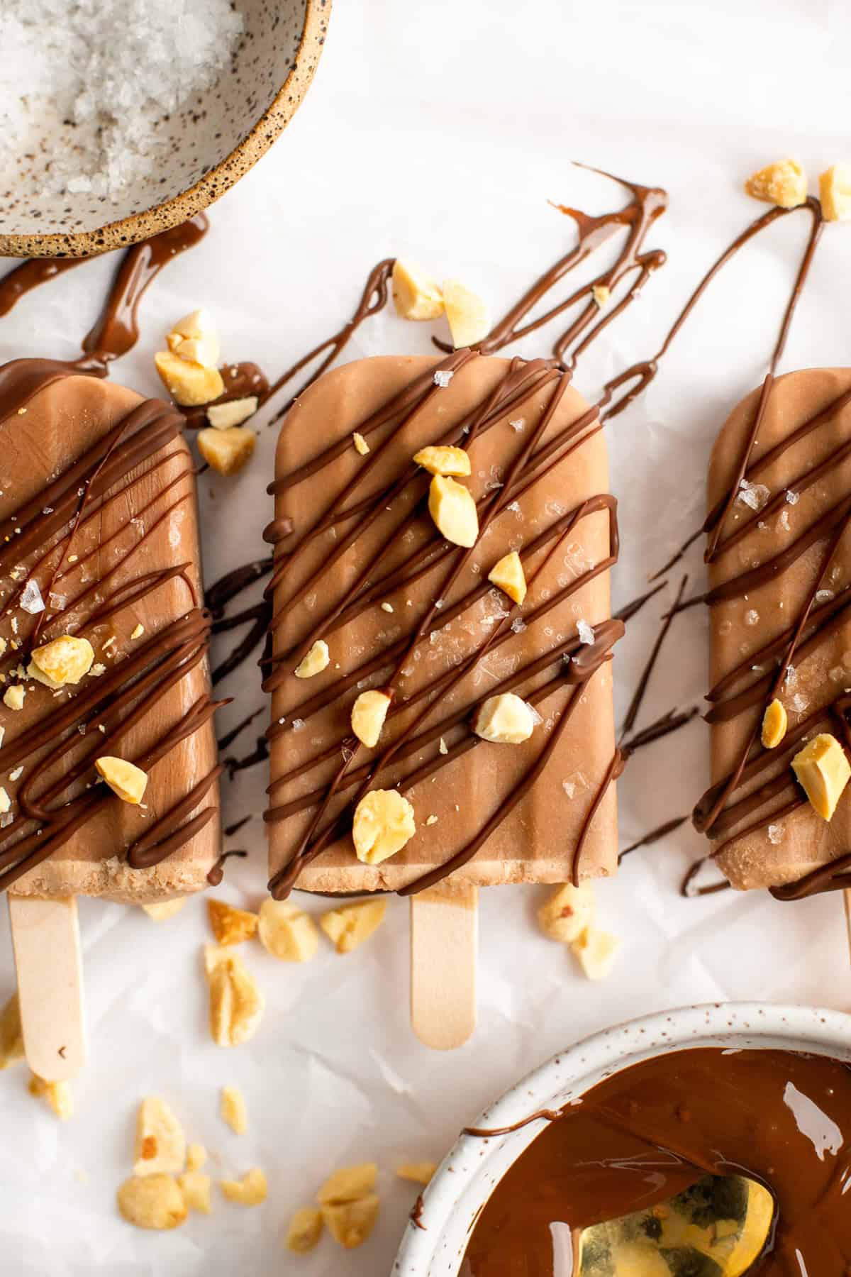 Three chocolate popsicles drizzled with dark chocolate and sprinkled with crushed nuts and sea salt are arranged on a parchment-covered surface.