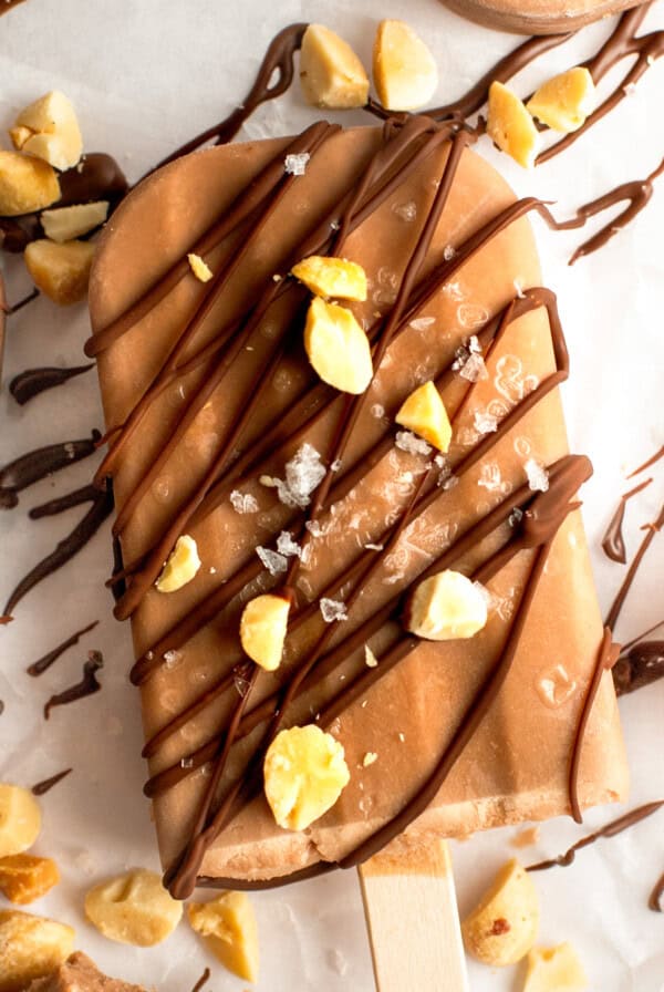 Chocolate-coated ice cream bar sprinkled with nuts and drizzled chocolate on parchment paper.