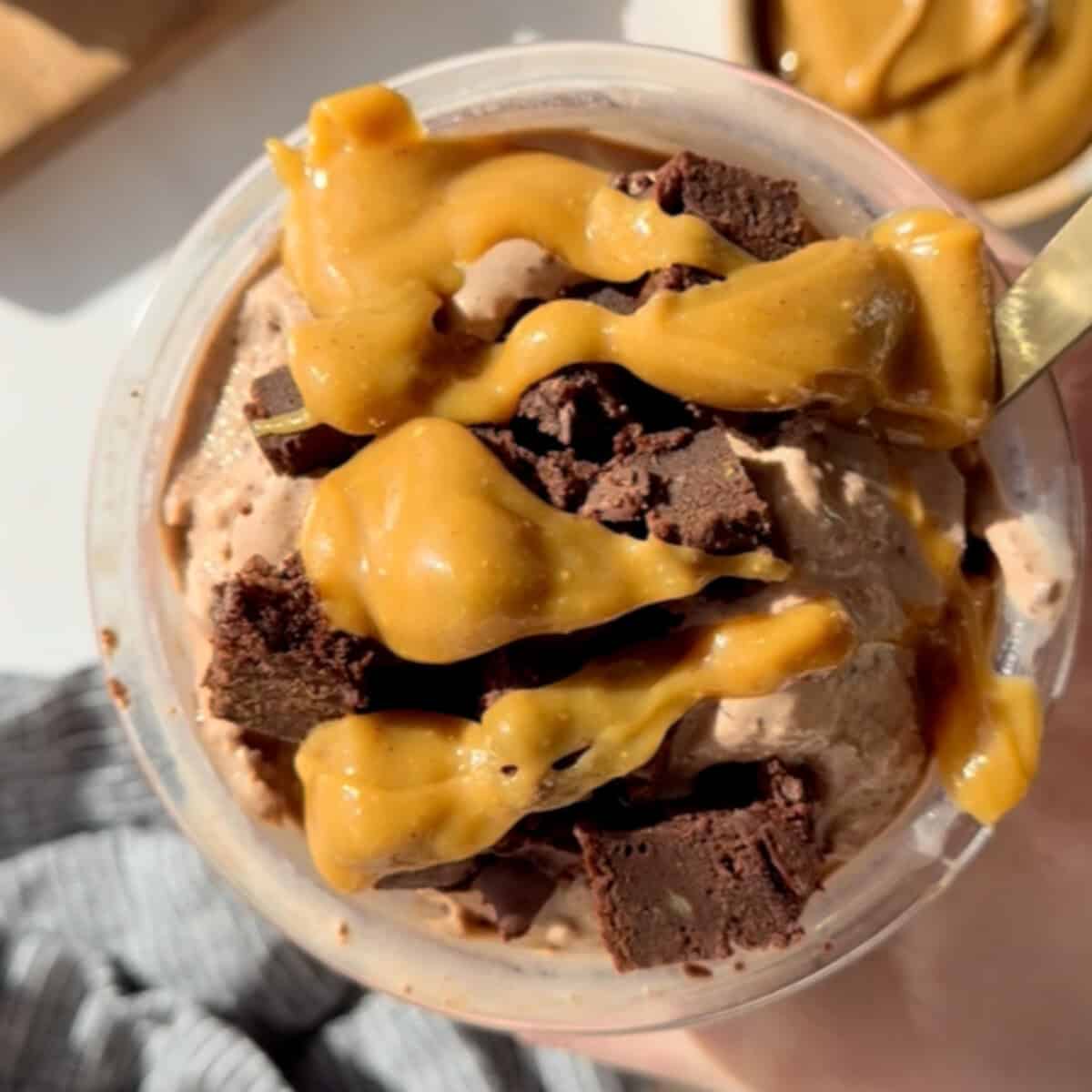 A bowl of chocolate ice cream topped with caramel sauce and chunks of chocolate brownies. A gold spoon is partially visible on the right.